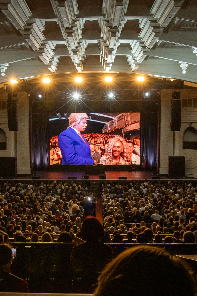 Large cinema screen displays performer to packed theatre audience beneath ornate geometric ceiling with golden lights.