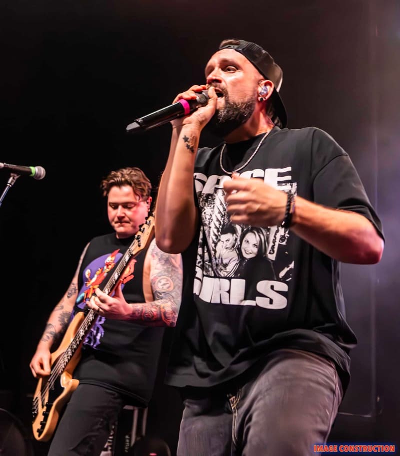 Bearded singer in black graphic tee performs at microphone with guitarist behind him on dark stage.