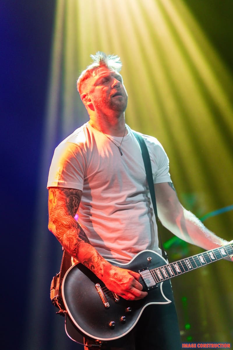 Guitarist with white spiky hair and tattooed arm performs on stage bathed in yellow and red spotlights.