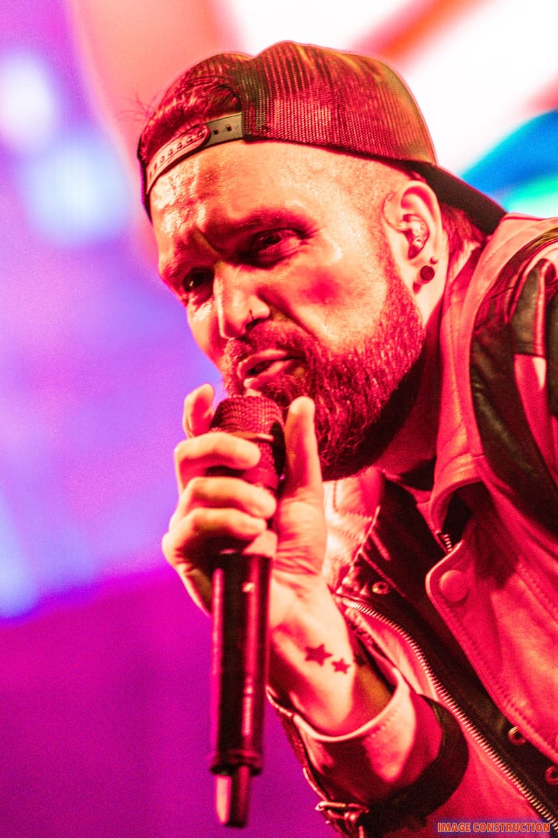 Bearded performer with red cap singing into microphone under vibrant pink and blue stage lighting.