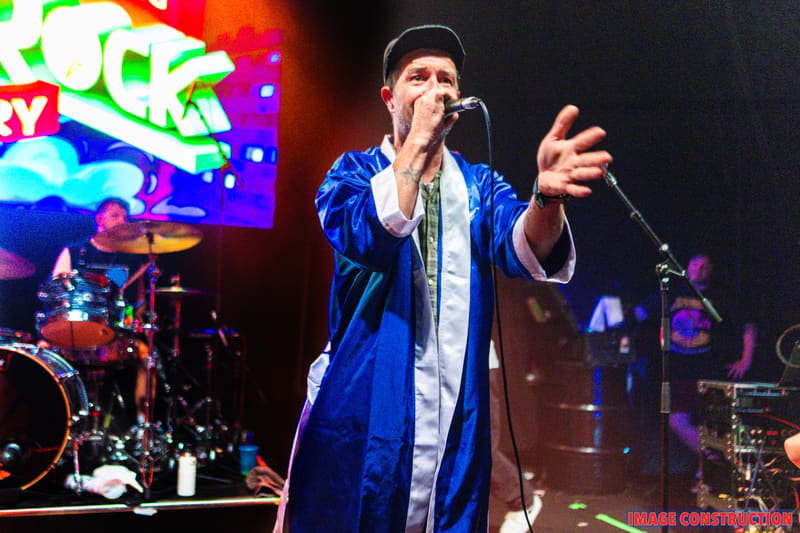 Performer in a blue satin jacket singing at a microphone with vibrant neon signage and band behind.