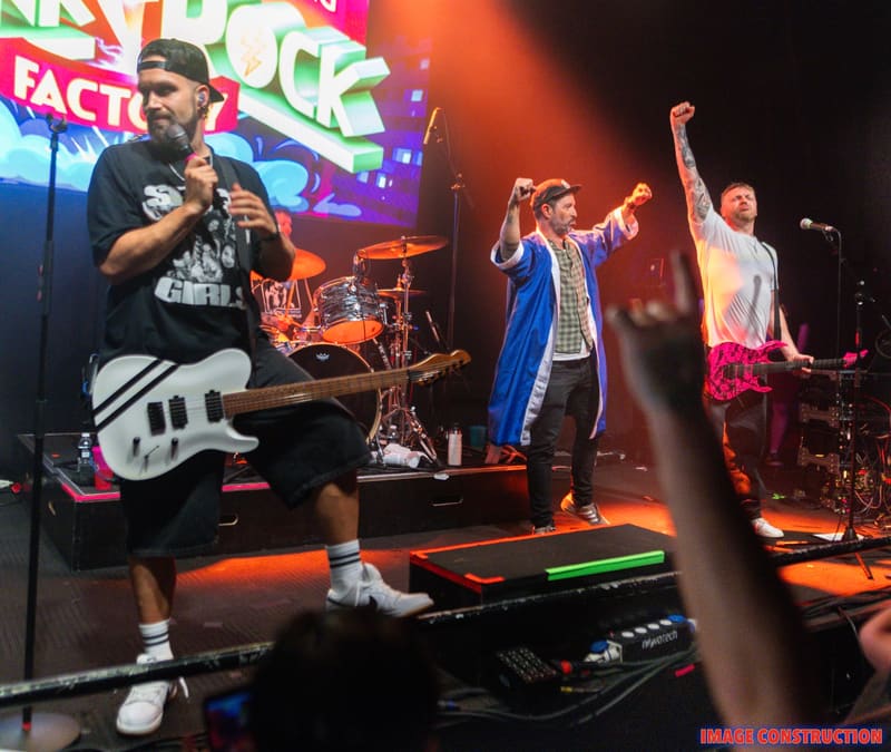 Band performing on stage with guitarist in foreground and three vocalists under colourful neon lighting at Punk Rock Factory.