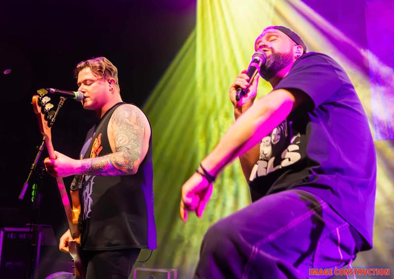 Two male performers sing into microphones under purple and yellow stage lighting during a punk rock concert.