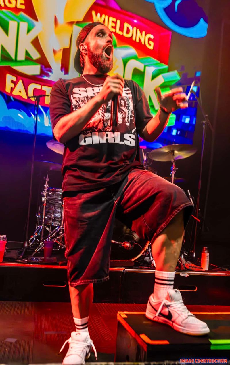 Performer in black graphic t-shirt and red shorts singing energetically on stage against vibrant neon signage backdrop.