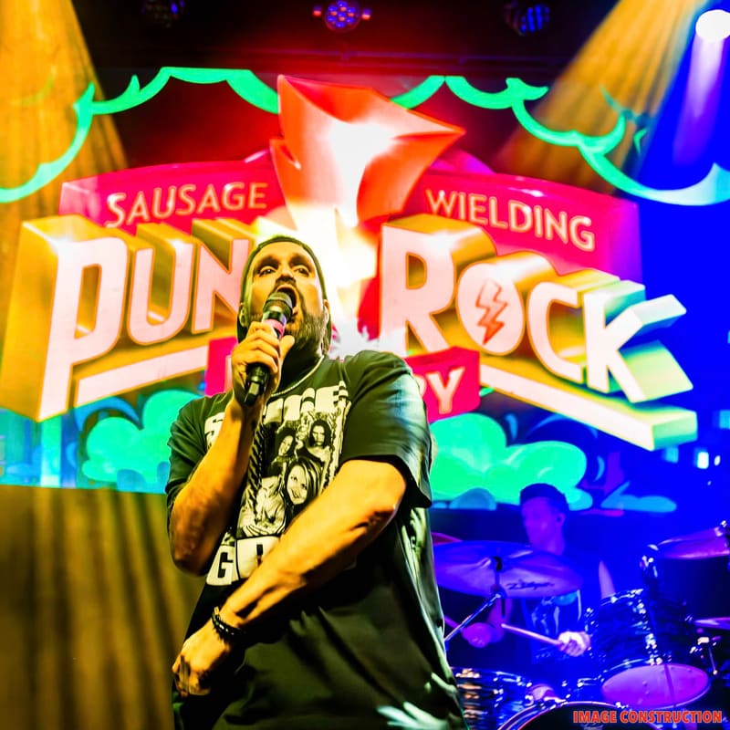 Singer performing at microphone with vibrant neon-coloured stage backdrop and band visible behind.