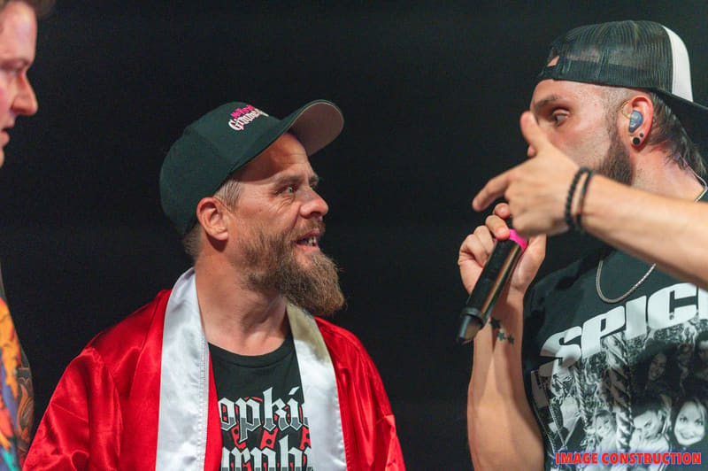 Two bearded men in baseball caps and casual wear engage in conversation on stage at Punk Rock Factory.