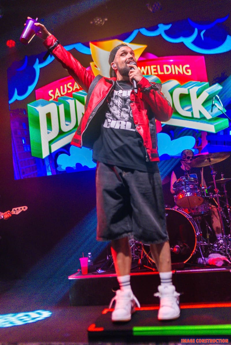 Performer in red jacket and black shorts raises fist on stage beneath vibrant neon signage, drummer visible in background.