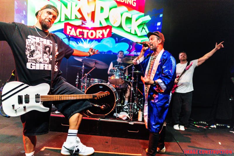 Guitarist in white bass and performer in blue jacket on stage beneath colourful Punk Rock Factory signage.