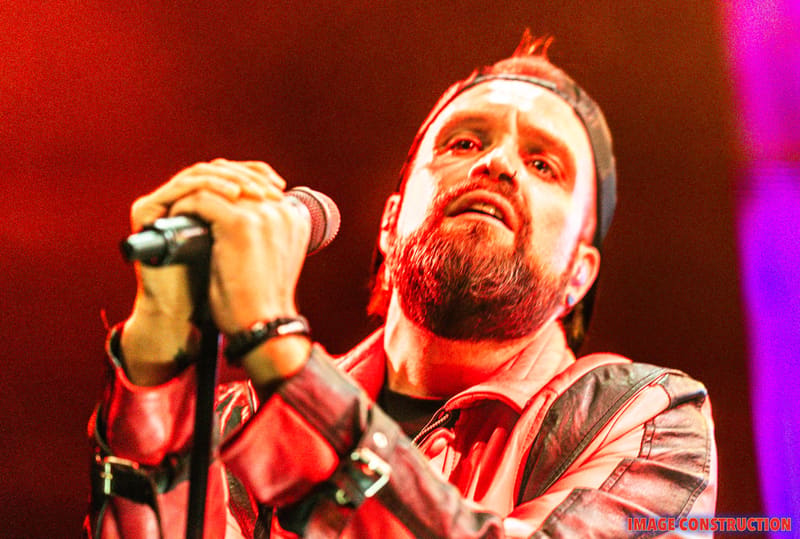 Bearded male vocalist singing into microphone under red and magenta stage lighting.