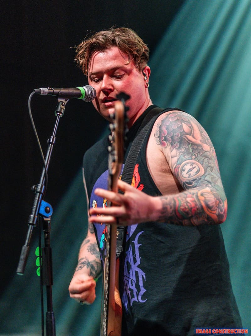 Tattooed musician performs at microphone with bass guitar against teal stage lighting.