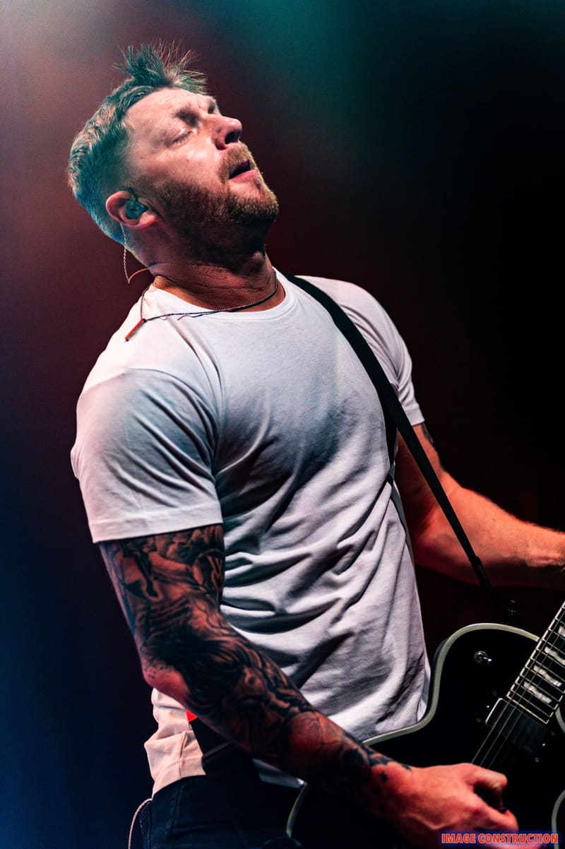 Bearded musician with tattooed arm playing guitar on stage, lit by red and blue stage lights.