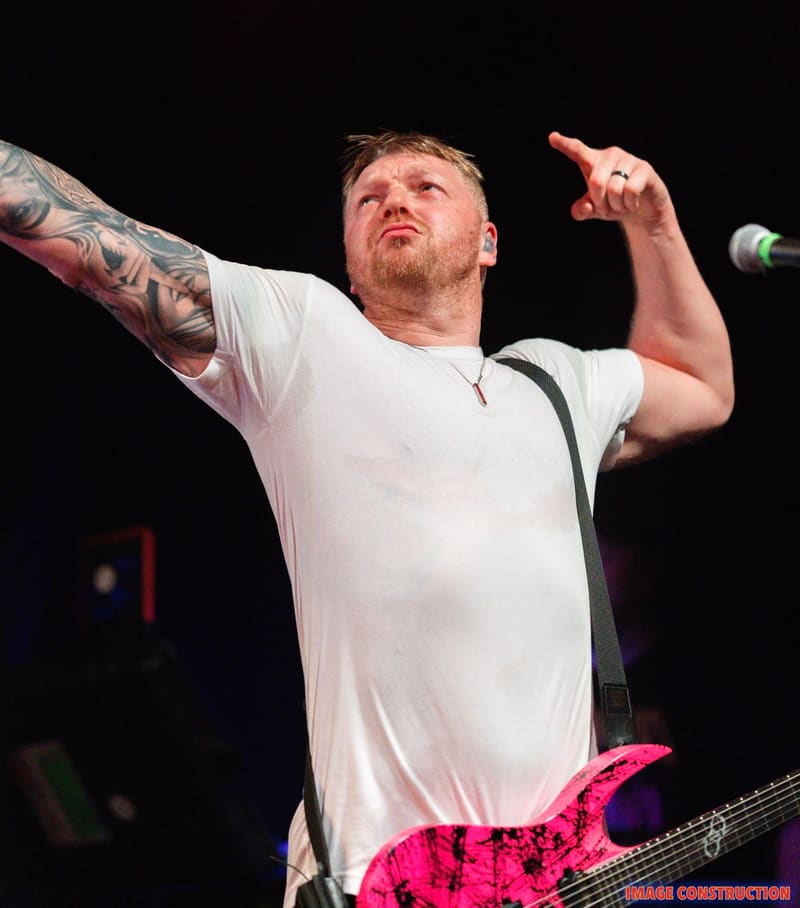 Musician performing with tattooed arm raised, holding a bright pink electric guitar against a dark stage background.