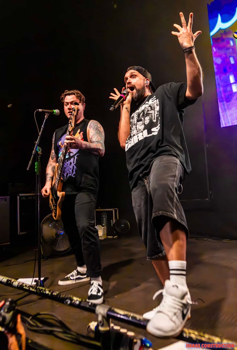Two musicians perform on stage at Punk Rock Factory, one playing guitar at a microphone while the other sings with raised arm, illuminated by purple stage lighting.