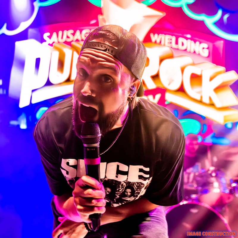 Performer in black headband and graphic t-shirt grips microphone against vibrant neon signage in pink and purple.