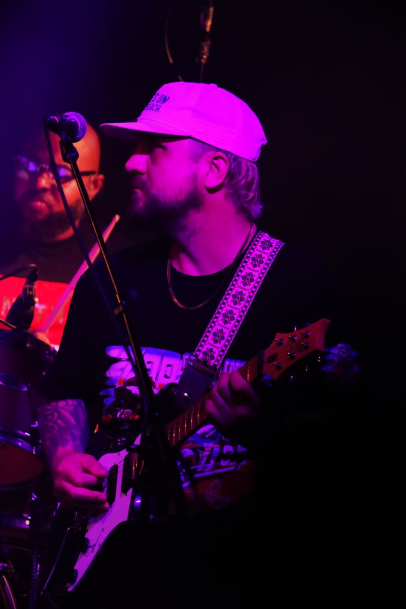 Guitarist performing at microphone under vibrant magenta and purple stage lighting.