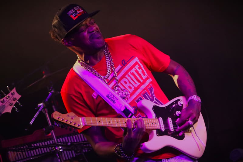 Guitarist in red shirt and cap playing electric guitar under purple and pink stage lighting.