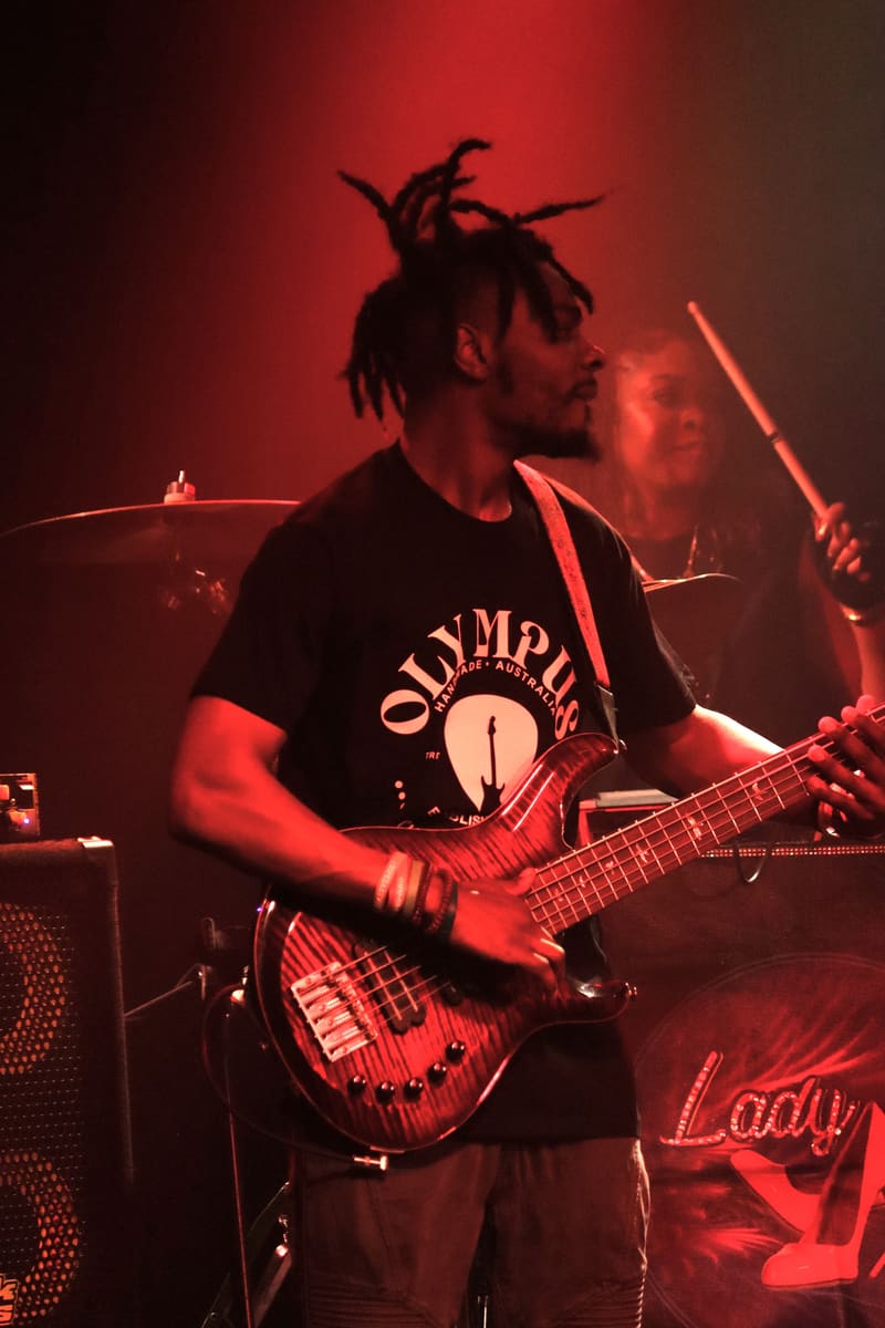 Guitarist with dreadlocks performing under red stage lighting, holding an electric guitar.