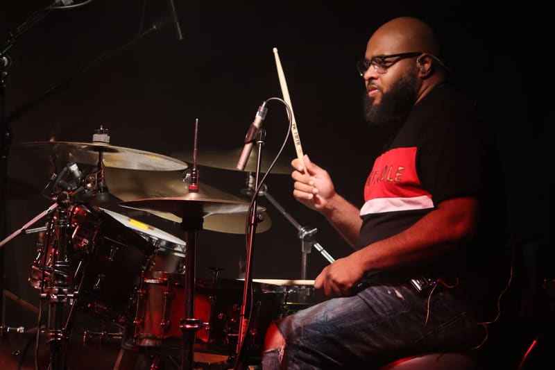 Drummer in red shirt performing at drum kit under stage lighting.
