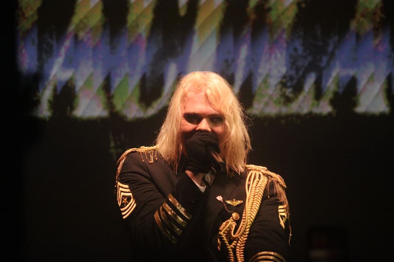 Performer with long white hair and full beard wearing an ornate gold-trimmed jacket under stage lighting.