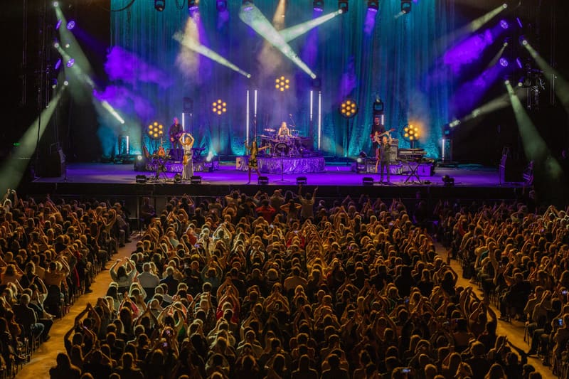 Large crowd watches performers on a purple-lit stage with suspended golden decorative elements during The Corrs concert.
