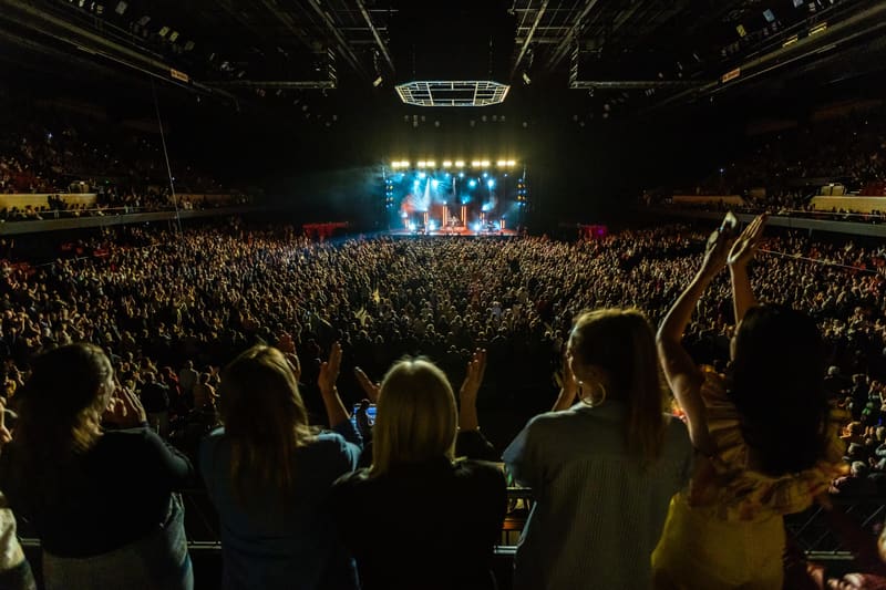 Large crowd with raised arms watching performers illuminated on stage at an indoor concert venue.