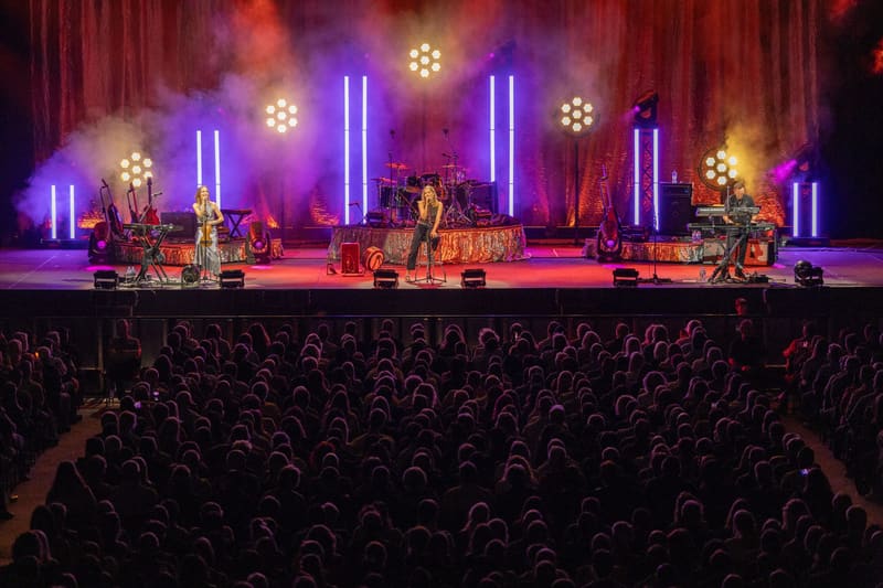 The Corrs perform on a stage lit by neon blue vertical lights, with a full audience visible in silhouette.