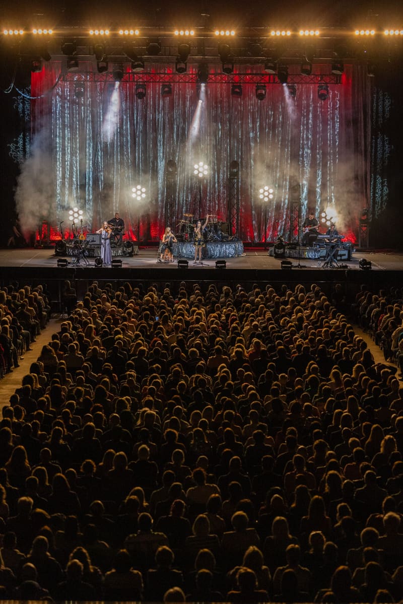 Band performing on stage with red lighting and dramatic curtains whilst audience watches from theatre seats below.