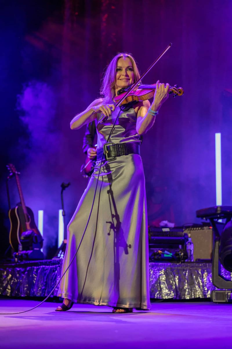 Violinist performing in a cream dress beneath purple stage lighting during The Corrs Australian Tour 2023.