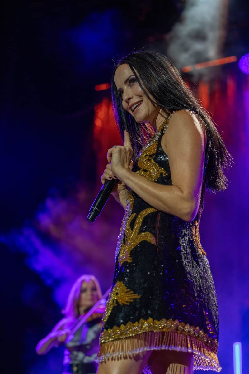 Singer performing at a microphone in a sequinned black and gold dress, illuminated by purple and blue stage lighting.