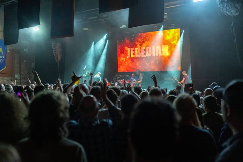 Crowd of concert-goers with raised hands facing a stage lit by blue spotlights and a large Jebediah logo screen.
