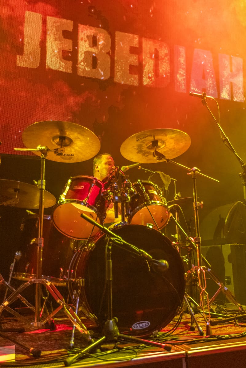 Drummer performing at drum kit on stage beneath Jebediah band name, bathed in warm orange and yellow stage lighting.