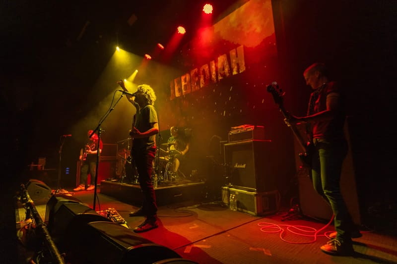Four-piece rock band Jebediah performing on stage bathed in warm orange and yellow stage lighting during their 2023 tour.