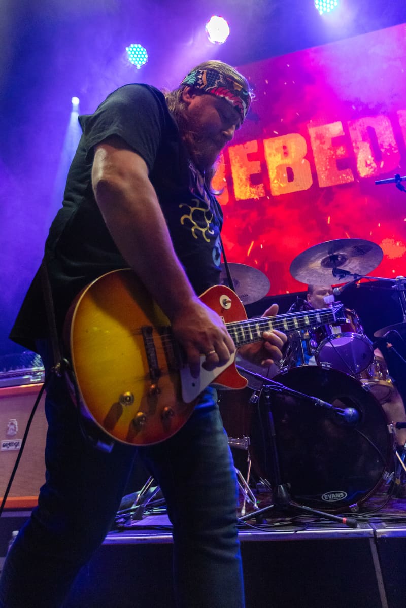 Guitarist performs with sunburst electric guitar under vibrant purple and pink stage lights during Jebediah's 2023 tour.
