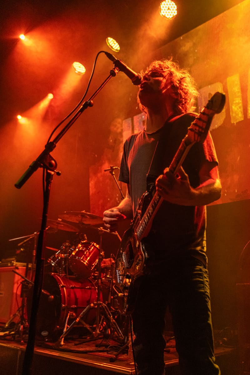 Musician performs with bass guitar on stage bathed in warm orange and red stage lighting during the Gum Up The Bearings tour.