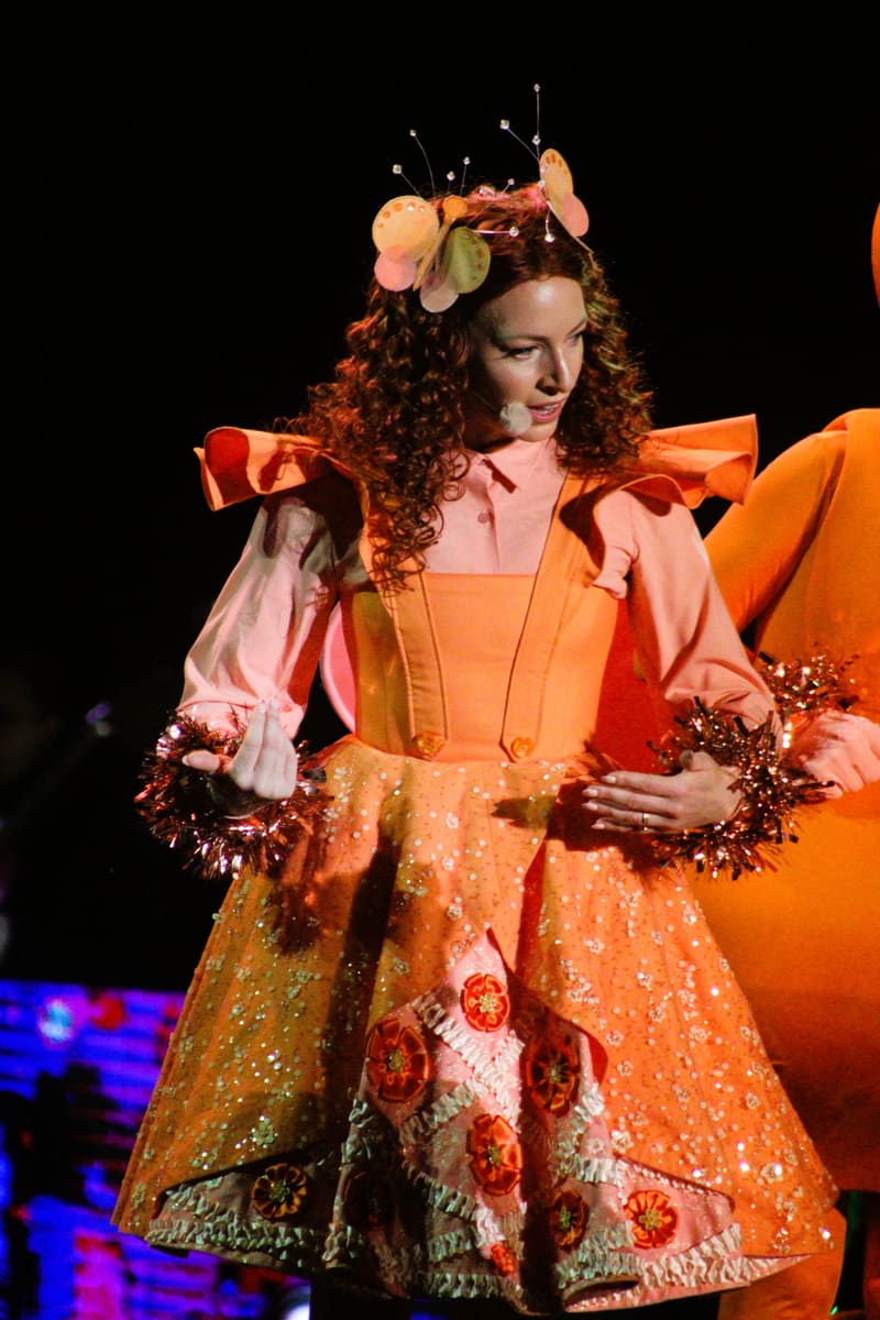 Performer in an ornate orange and pink costume with tinsel garland and decorative headpiece stands on stage during Adelaide Carols By Candlelight.