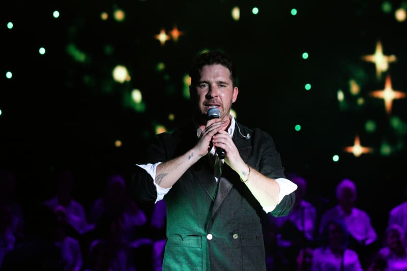 Man singing into microphone against festive green and gold starlit backdrop at Adelaide Carols By Candlelight.