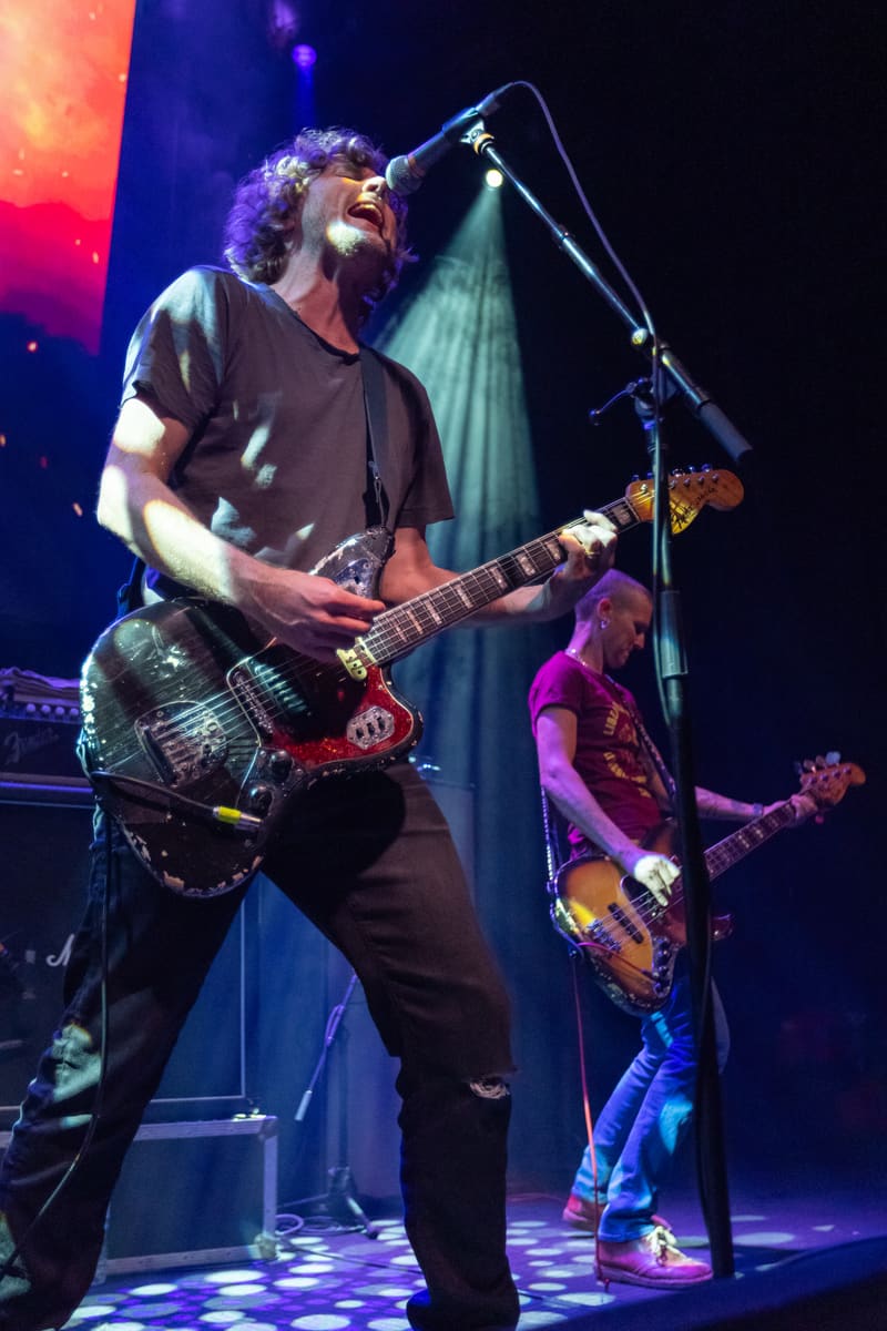 Guitarist with curly hair singing into microphone whilst playing electric guitar on stage, with band members and coloured stage lighting visible behind.