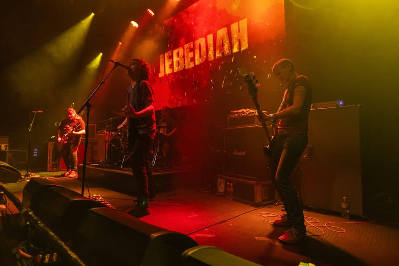 Band members perform on stage bathed in red and yellow stage lighting, with "JEBEDIAH" projected behind them.
