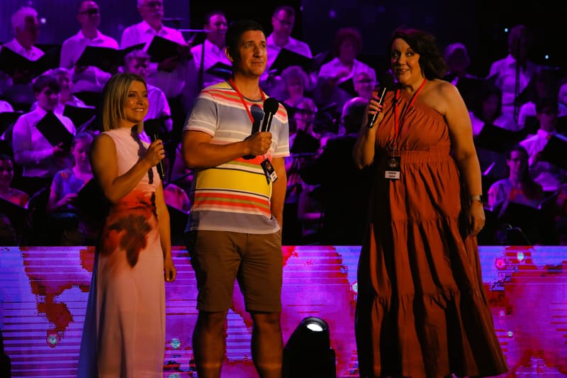 Three performers stand centre stage under purple lighting during Adelaide Carols By Candlelight, with audience members visible in the background.