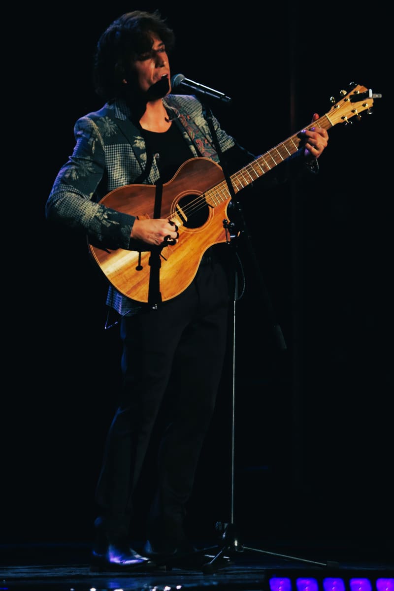 Musician playing acoustic guitar and singing at microphone on dark stage, lit by blue spotlights.