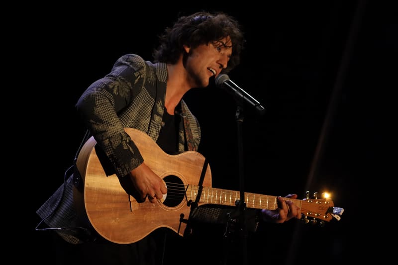 Musician with curly hair performing with an acoustic guitar under stage lighting at Adelaide Carols By Candlelight.
