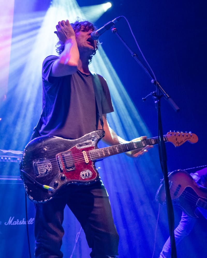 Musician performs with red electric bass guitar at microphone under blue stage lighting during Jebediah's Gum Up The Bearings tour.