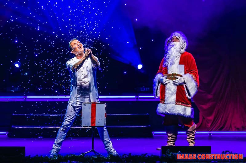 Performer in light-coloured top singing on stage beside a figure dressed as Santa Claus, both lit by blue and purple stage lighting.