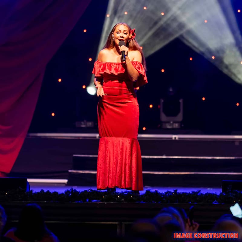 Performer in a red sequined gown singing at a microphone on a stage lit by spotlights during The Christmas Spectacular.