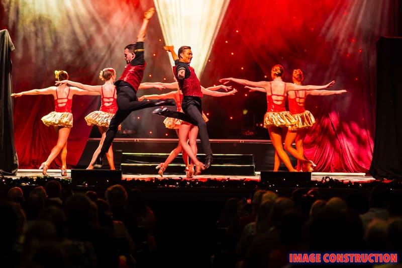 Dancers in red and gold costumes perform synchronized moves on stage under warm red lighting during The Christmas Spectacular.