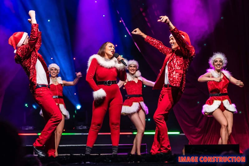 Five performers in red sequined costumes dance on stage under purple and blue spotlights during The Christmas Spectacular.