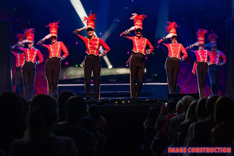 Performers in matching red and gold military-style jackets stand on stage under warm spotlights during The Christmas Spectacular.