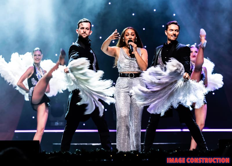 Three performers in sequined outfits dance on stage amid swirling white fabric and blue lighting during The Christmas Spectacular.
