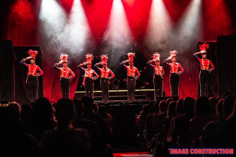 Seven performers in red sequined outfits stand on stage beneath dramatic red and white spotlights during The Christmas Spectacular.