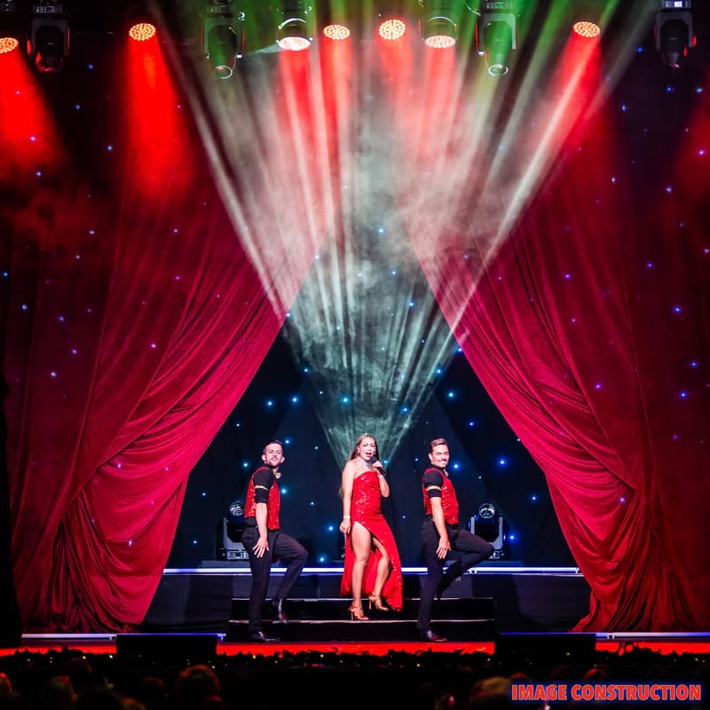 Three performers in red costumes stand centre stage beneath dramatic red and white light beams, flanked by velvet curtains.
